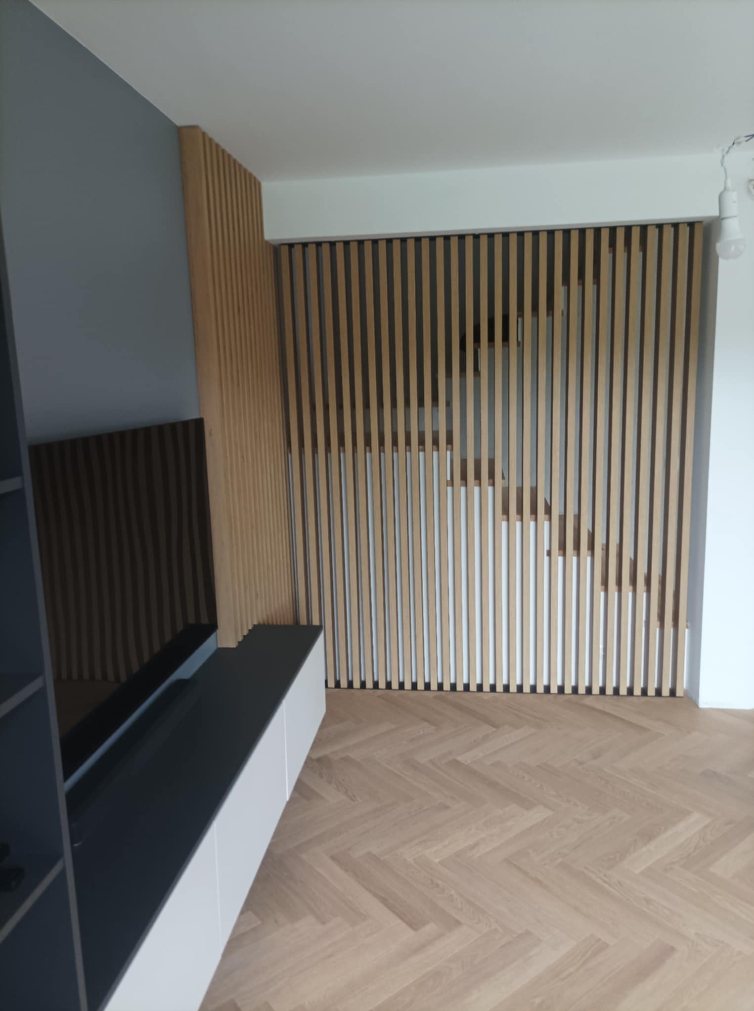 Nowoczesny salon z lamelami drewnianymi na ścianie, szafką RTV z białymi frontami i parkietem w jodełkę. Minimalistyczny design, jasne wnętrze.