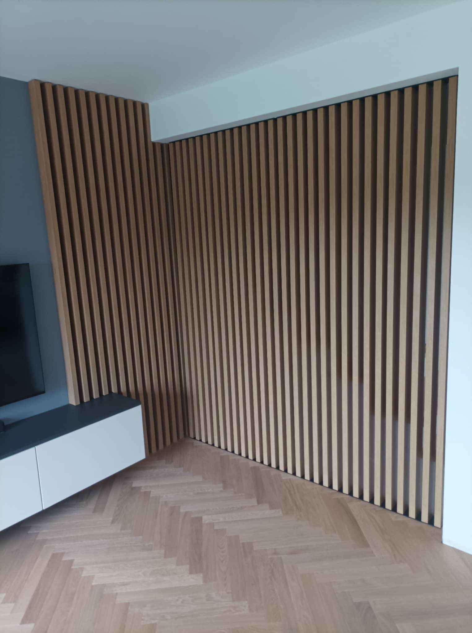 Drewniane lamele ścienne w jasnym odcieniu, tworzące dekoracyjny panel w nowoczesnym wnętrzu. Widoczny fragment szafki RTV i parkiet w jodełkę.