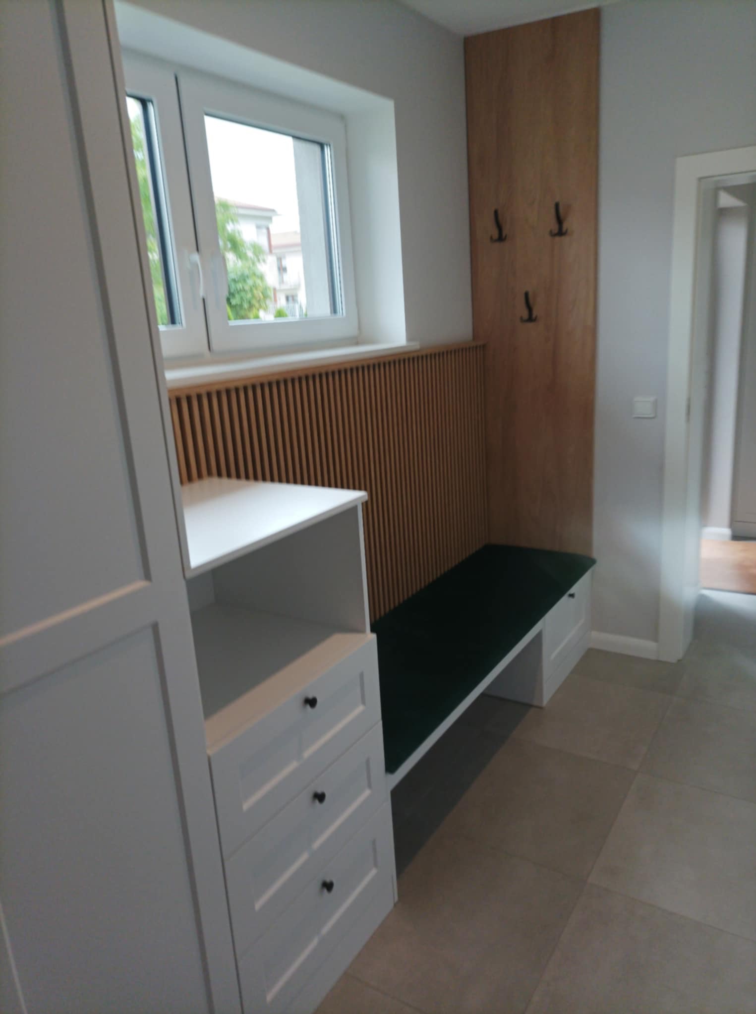 Funkcjonalna zabudowa przedpokoju z szafką, siedziskiem z zielonym obiciem i drewnianym panelem z wieszakami. Jasne wnętrze z oknem i szarą podłogą.