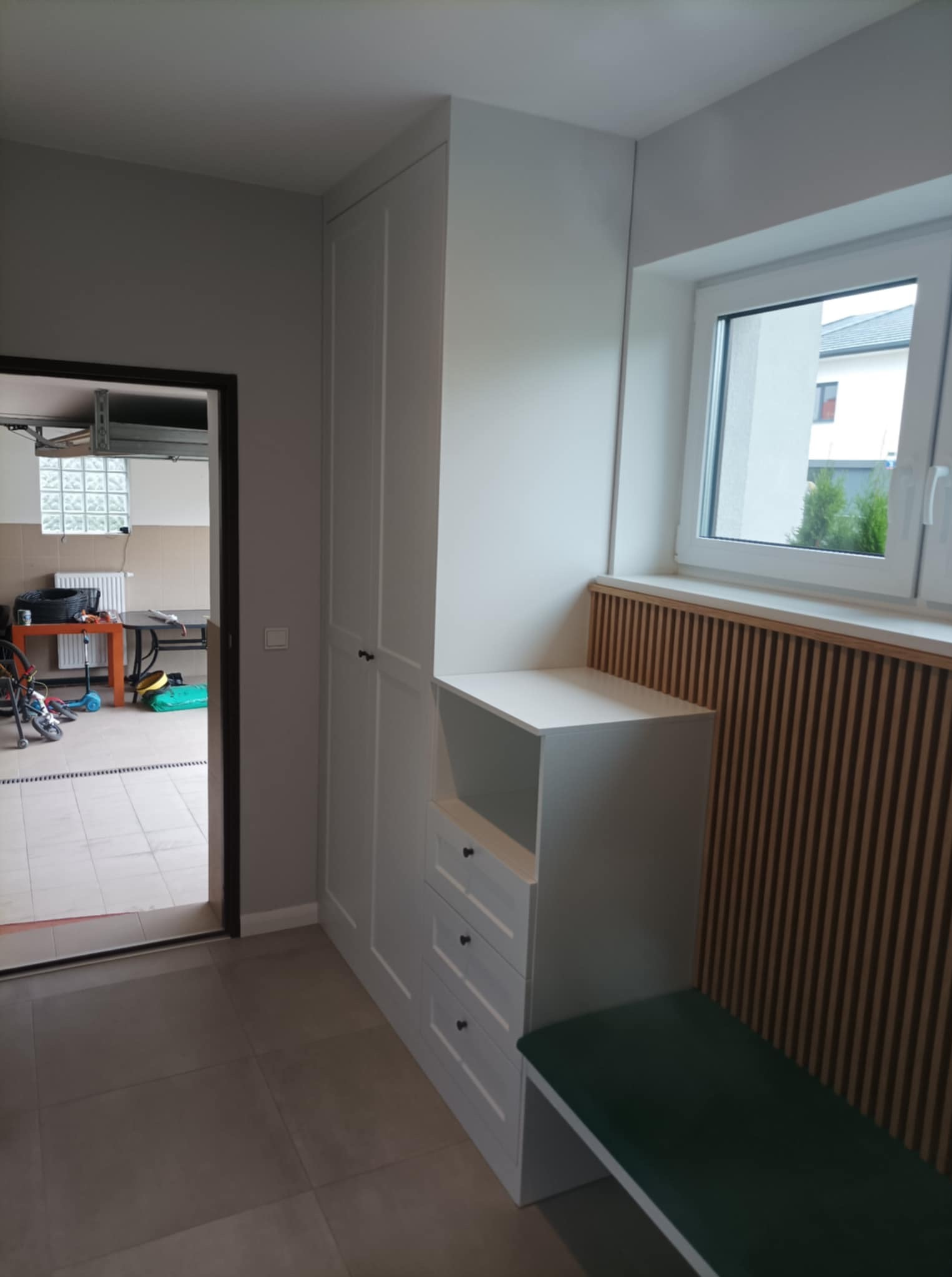 Biała szafa wnękowa z siedziskiem i szufladami, dopasowana do wnęki obok okna. Widoczna drewniana boazeria i fragment garażu w tle.