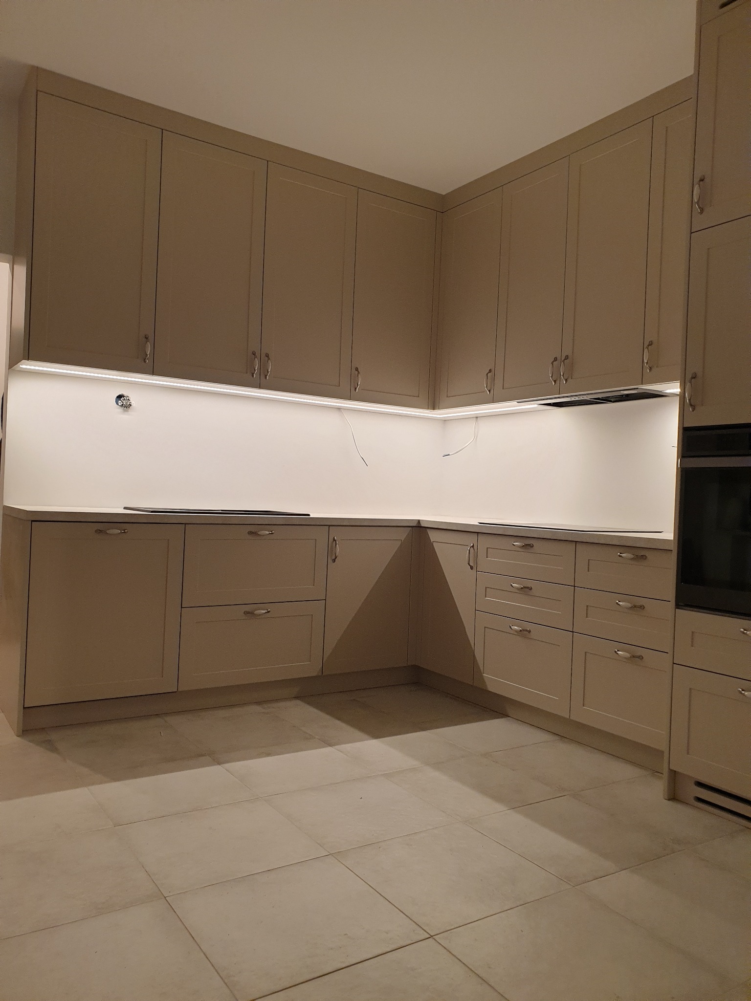 Jasna, narożna zabudowa kuchenna z górnymi szafkami do sufitu, podświetleniem LED pod szafkami i jasnymi płytkami na podłodze.