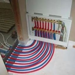 Rozdzielacz ogrzewania podłogowego z rurami czerwono-niebieskimi układanymi w łuk na białym styropianie, widoczne przewody i elementy instalacji wewnątrz szafki.