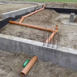 Fundamenty budynku z ułożonymi rurami kanalizacyjnymi w wykopie, widoczne zbrojenia i butelka wody obok rury.