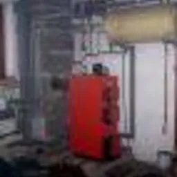 Czerwona skrzynka elektryczna z bezpiecznikami, zamontowana w pomieszczeniu technicznym z widocznymi rurami i elementami instalacji.