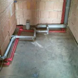 Surowe wnętrze z ceglanymi ścianami w trakcie instalacji hydraulicznej: szare i czerwone rury ułożone na betonowej podłodze, widoczne przyłącza wodne i kanalizacyjne.