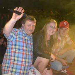 Wokalista z podniesionym mikrofonem i uśmiechnięta blondynka w skórzanej kurtce oraz mężczyzna w czerwonej czapce na tle publiczności i sceny oświetlonej reflektorami.