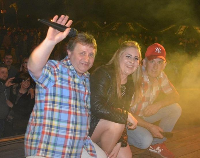 Wokalista z podniesionym mikrofonem i uśmiechnięta blondynka w skórzanej kurtce oraz mężczyzna w czerwonej czapce na tle publiczności i sceny oświetlonej reflektorami.