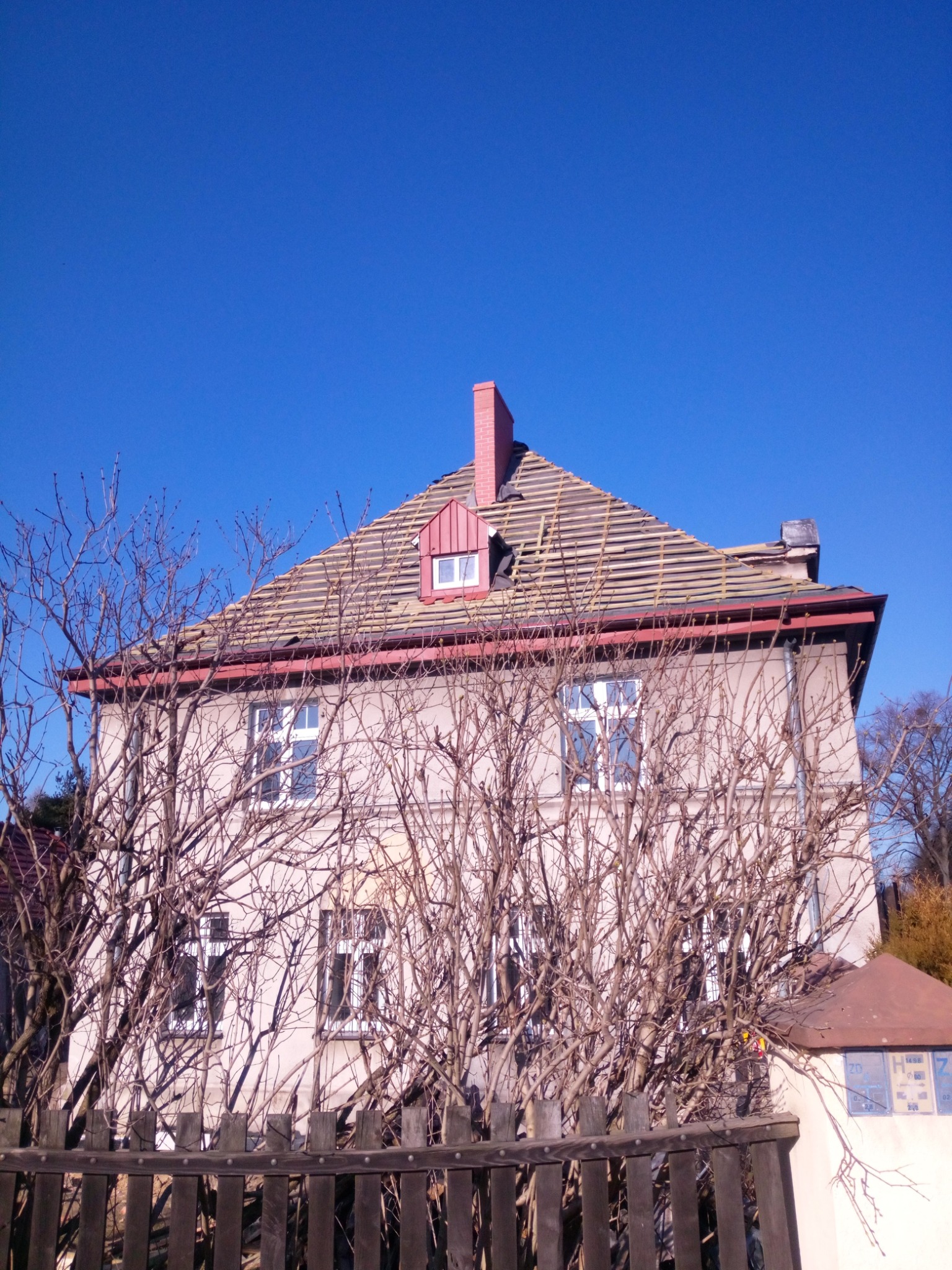 Dom z częściowo zdjętym dachem, widoczne łaty pod dachówką, komin z czerwonej cegły, okno dachowe, ujęcie przez gałęzie drzew.