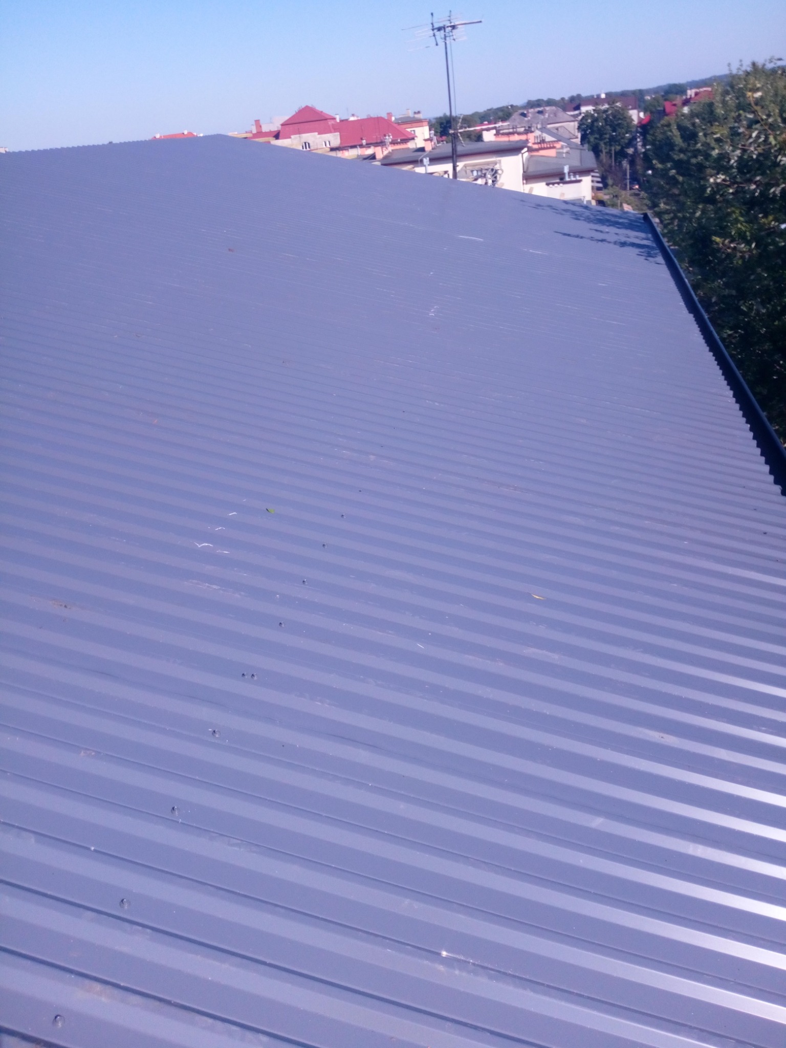 Nowy, szary dach z blachy falistej, widok z góry, w tle budynki mieszkalne i drzewa.