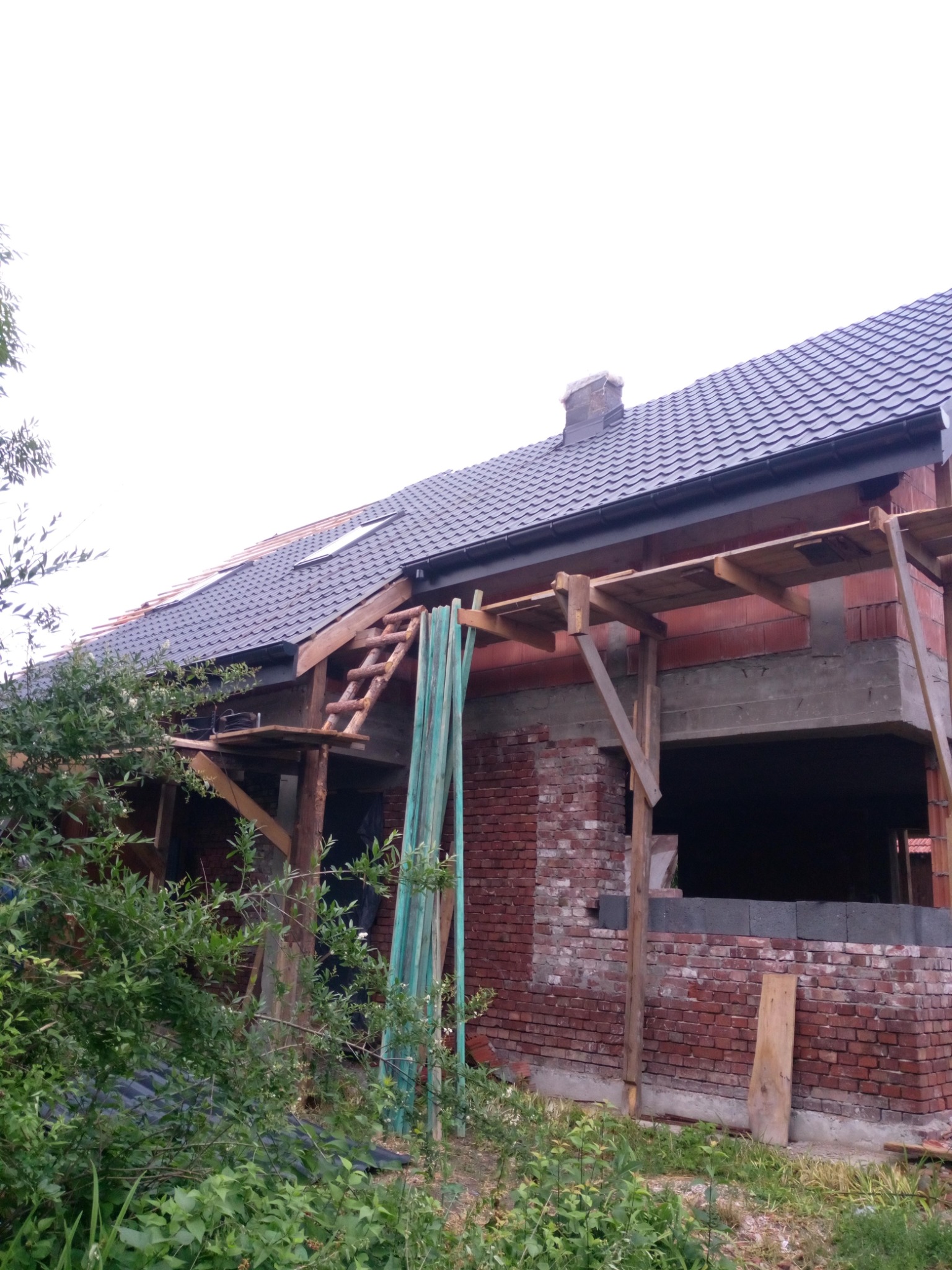 Dom w trakcie budowy z częściowo położonym ciemnoszarym dachem, widoczne rusztowania i ślady prac murarskich na ścianach.