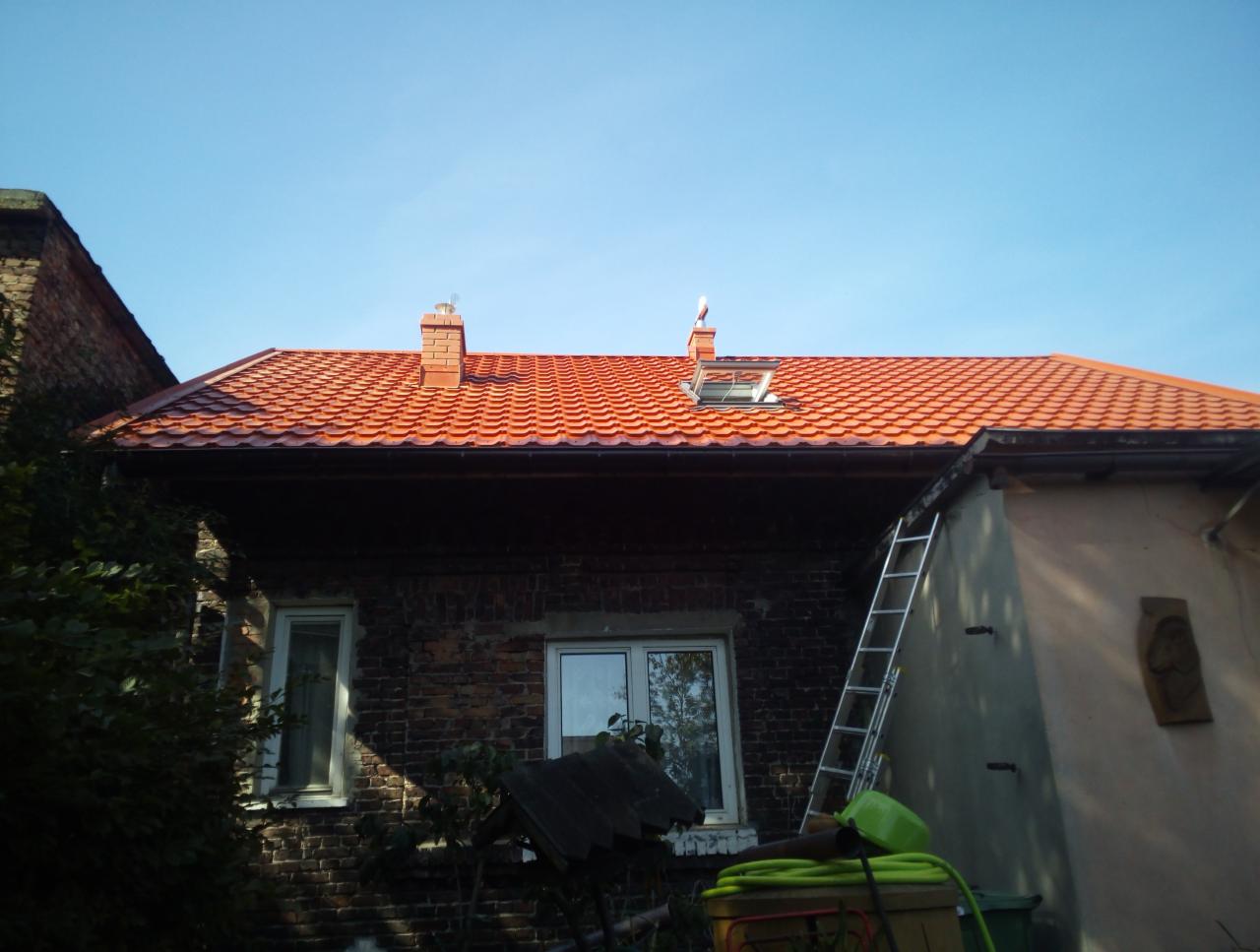 Ceglany dom z nową, czerwoną dachówką karpiówką, kominami i oknem dachowym, drabina oparta o ścianę, bocianie gniazdo na jednym z kominów.
