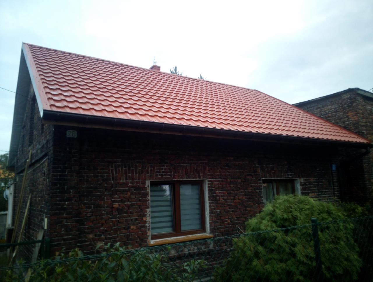 Dom z ciemnej cegły z nowym, błyszczącym, czerwonym dachem z blachodachówki modułowej i nowymi rynnami.