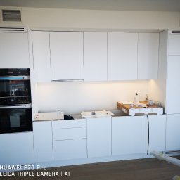 TIMWEK KATARZYNA KORBUT - Nowoczesna, biała zabudowa kuchenna z wbudowanym piekarnikiem Miele, widoczna w trakcie montażu, z elementami opakowań i narzędzi na blacie.