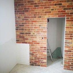 TIMWEK KATARZYNA KORBUT - Ściana z cegły klinkierowej w trakcie montażu, widoczny fragment ściany przygotowany pod okładzinę, drabina w przejściu, żółta szpachelka na podłodze.