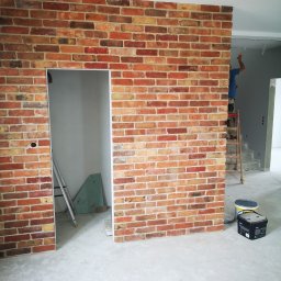 TIMWEK KATARZYNA KORBUT - Ściana z cegieł klinkierowych z otworem drzwiowym, widoczna drabina i malarz w tle, surowy beton na podłodze, pomieszczenie w trakcie wykańczania.