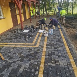 TIMWEK KATARZYNA KORBUT - Ekipa brukarzy układa kostkę brukową w geometryczny wzór z żółtymi liniami na zewnątrz budynku z drewnianym balkonem, widoczne narzędzia i materiały budowlane.