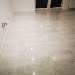 TIMWEK KATARZYNA KORBUT - Jasne, błyszczące płytki podłogowe imitujące marmur w pustym pokoju, widoczne wystające rury instalacji wodnej przy ścianie.