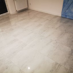 TIMWEK KATARZYNA KORBUT - Jasne, polerowane płytki podłogowe z szarym wzorem żyłkowania, widoczny fragment niebieskich drzwi przesuwnych w stylu stodoły oraz biały grzejnik.