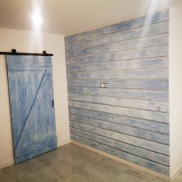 TIMWEK KATARZYNA KORBUT - Niebieska, przesuwna brama stodoły na czarnym torze obok ściany z desek pomalowanych na niebiesko-biało, z widocznymi gniazdkami elektrycznymi i drzwiami wejściowymi w tle.