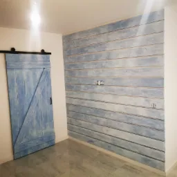 Niebieska, przesuwna brama stodoły na czarnym torze obok ściany z desek pomalowanych na niebiesko-biało, z widocznymi gniazdkami elektrycznymi i drzwiami wejściowymi w tle.