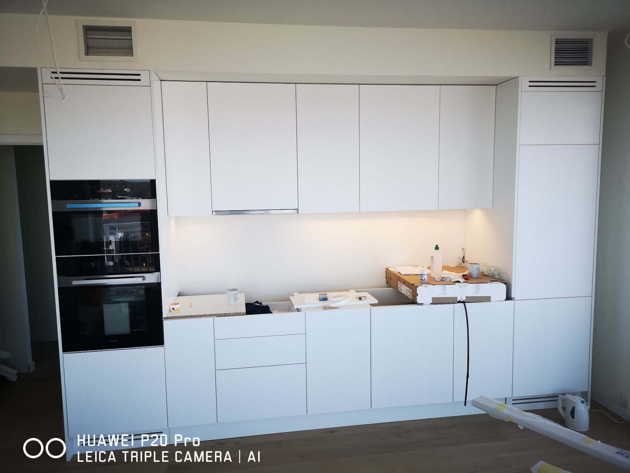 Nowoczesna, biała zabudowa kuchenna z wbudowanym piekarnikiem Miele, widoczna w trakcie montażu, z elementami opakowań i narzędzi na blacie.