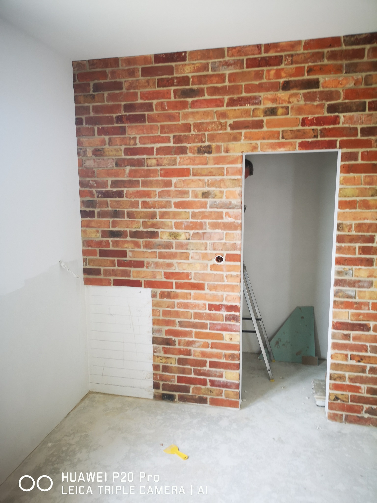 Ściana z cegły klinkierowej w trakcie montażu, widoczny fragment ściany przygotowany pod okładzinę, drabina w przejściu, żółta szpachelka na podłodze.