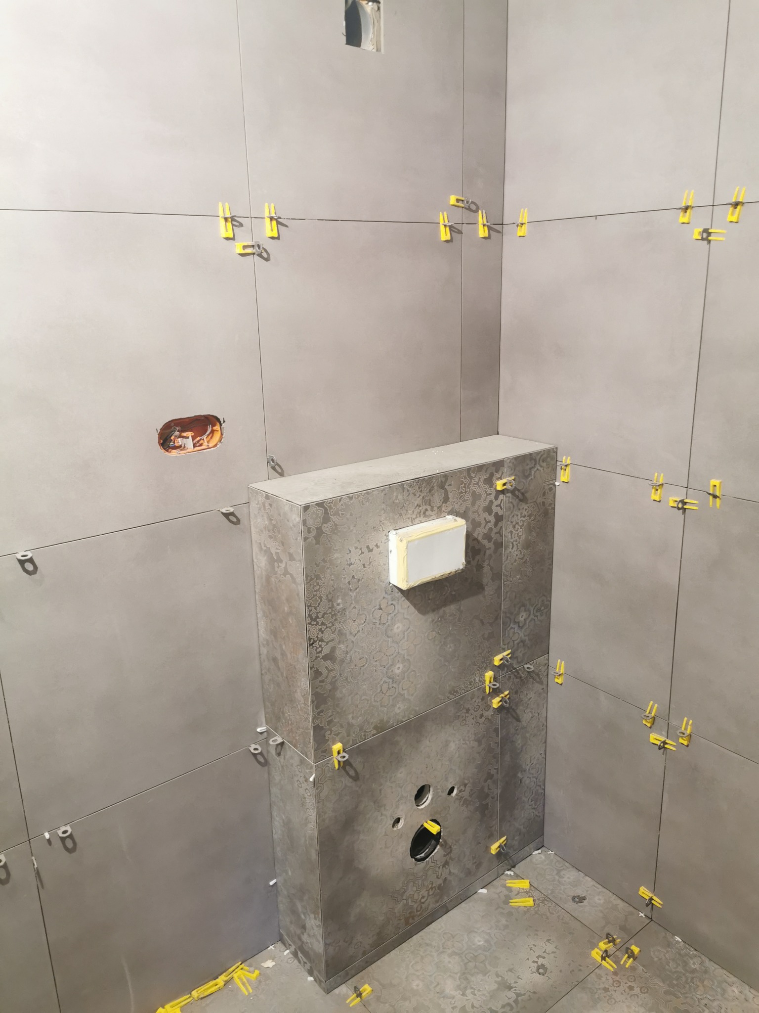 Szare płytki ceramiczne w trakcie montażu w łazience, widoczne żółte klipsy poziomujące, otwór na gniazdko elektryczne i zabudowa stelaża WC z wzorzystymi płytkami.