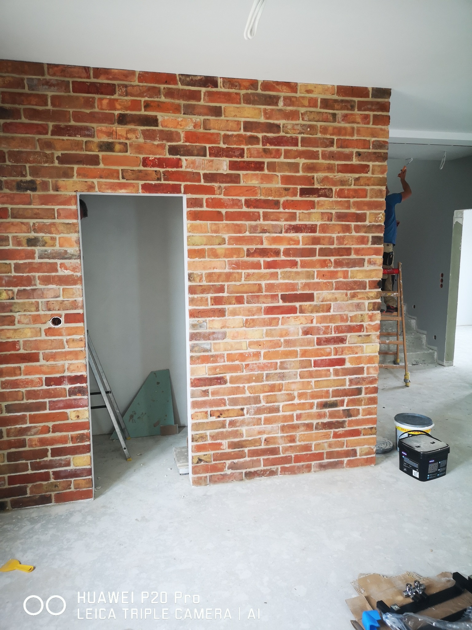 Ściana z cegieł klinkierowych z otworem drzwiowym, widoczna drabina i malarz w tle, surowy beton na podłodze, pomieszczenie w trakcie wykańczania.