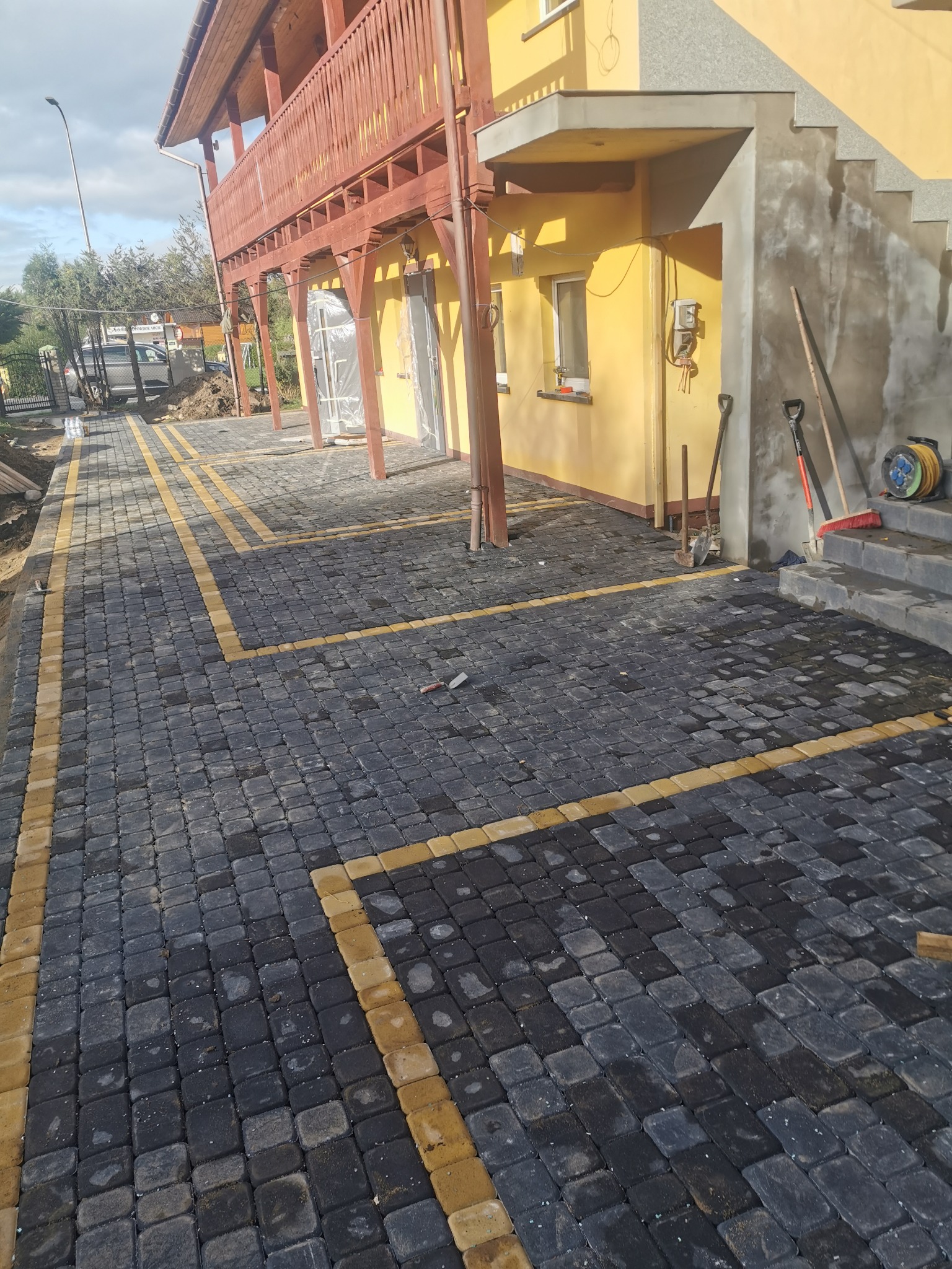 Ułożona kostka brukowa w kolorze grafitowym z żółtymi obramowaniami, widoczne narzędzia budowlane przy ścianie budynku.