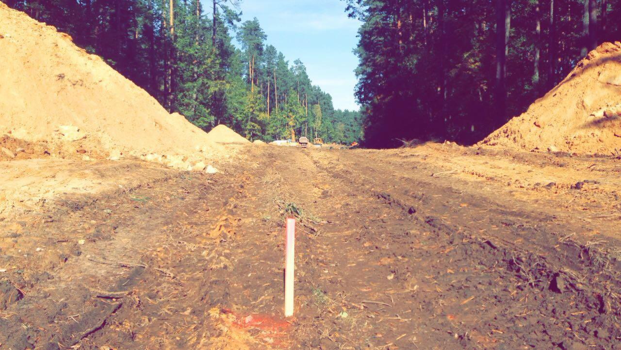 Teren budowy z wytyczonym obszarem, widoczne hałdy ziemi i las w tle, pomarańczowy słupek geodezyjny wbity w ziemię.