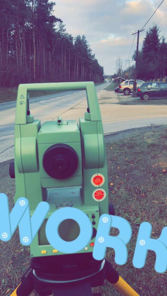 Zielony teodolit ustawiony przy drodze, w tle las i zaparkowane samochody. Na zdjęciu nałożony napis 'WORK'.