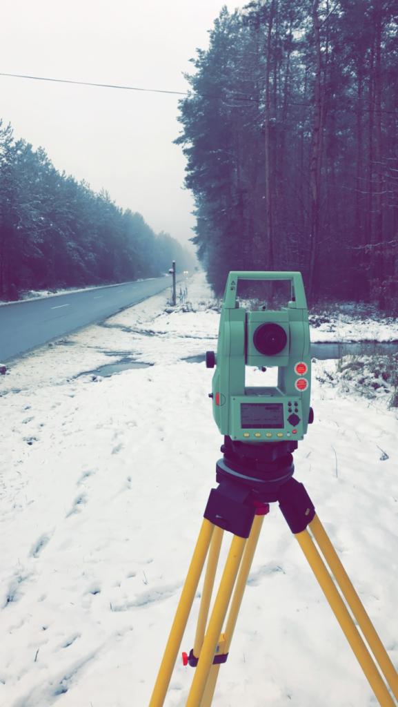 Zielony teodolit ustawiony na żółtym statywie w zasypanym śniegiem terenie, w tle droga i las we mgle.
