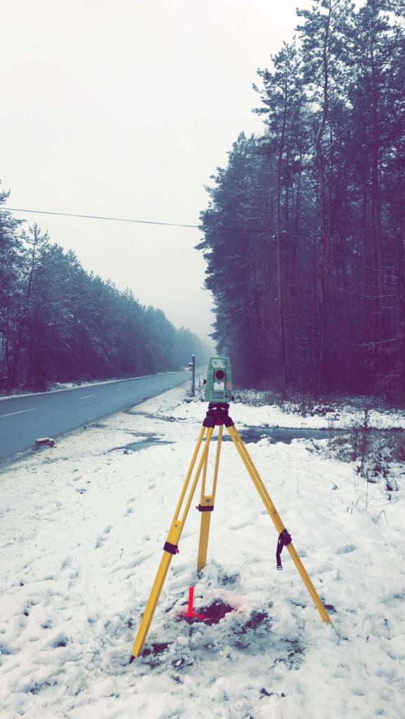 Pomiar geodezyjny przy drodze w zimowej scenerii: teodolit na żółtym statywie ustawiony na śniegu, w tle droga i zasypany śniegiem las.