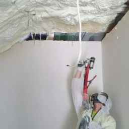 Pracownik w kombinezonie ochronnym aplikuje piankę PUR na poddasze z widocznymi fragmentami niezabezpieczonych elementów konstrukcyjnych dachu.