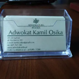 Wizytówka adwokata Kamila Osiki w przezroczystym podajniku, z adresem, numerem telefonu, adresem e-mail i godzinami otwarcia, umieszczona na drewnianym biurku.