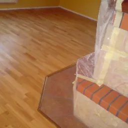 Jasna, odnowiona podłoga parkietowa sąsiaduje z zabezpieczonym folią i taśmą malarską fragmentem kominka z cegieł.