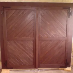 Dwie drewniane okiennice z ukośnym wzorem desek, zawieszone na ciemnych zawiasach, gotowe do montażu.