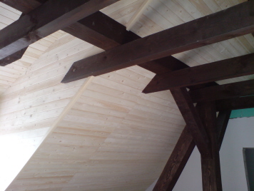 Wnętrze poddasza z widoczną konstrukcją drewnianą: jasne deski boazeryjne pokrywające skośną ścianę i sufit, ciemne, lakierowane belki stropowe, fragment ściany pomalowanej na biało oraz zielona...