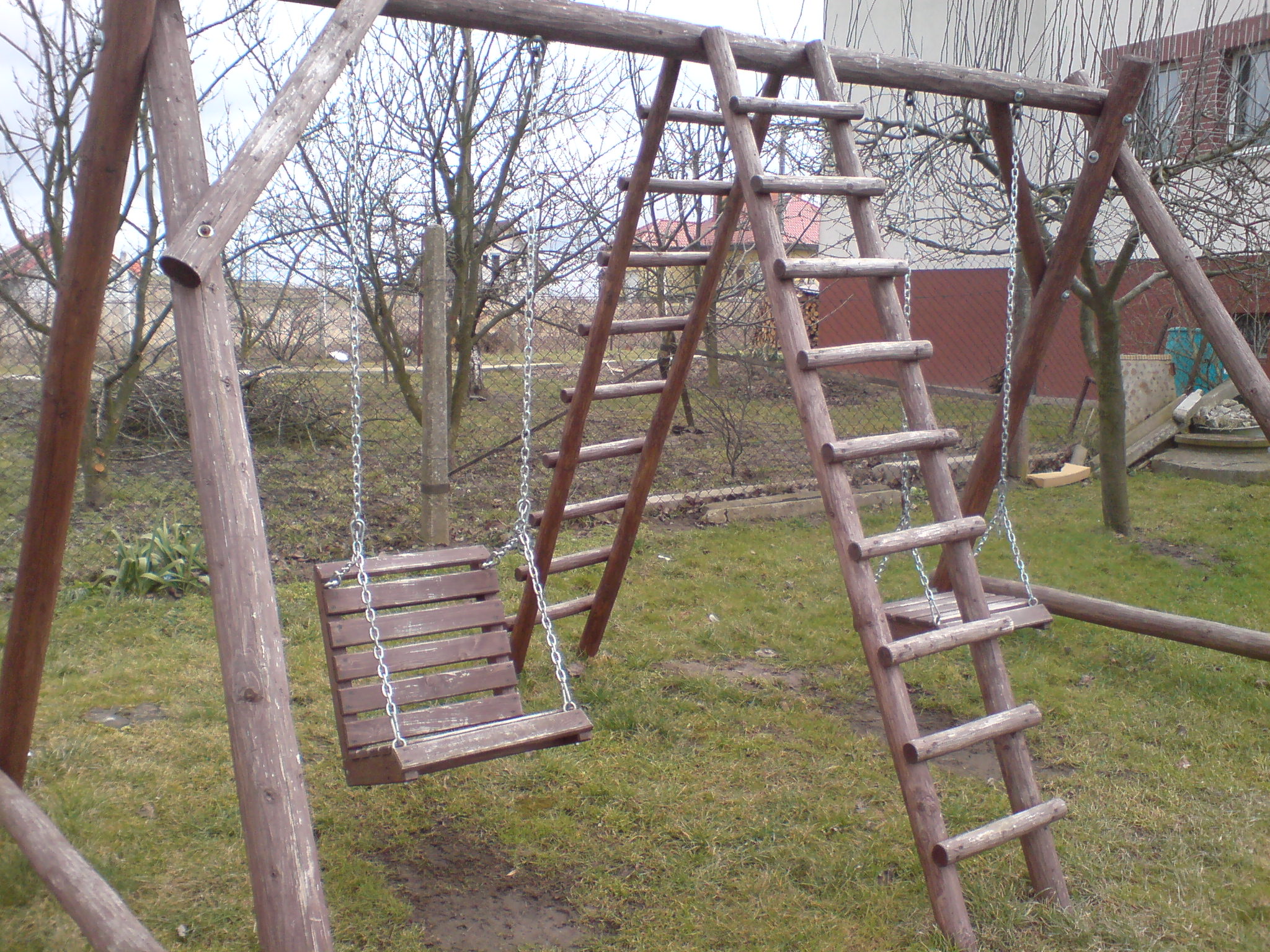 Drewniany plac zabaw z huśtawką i drabinką na tle ogrodu z drzewami.