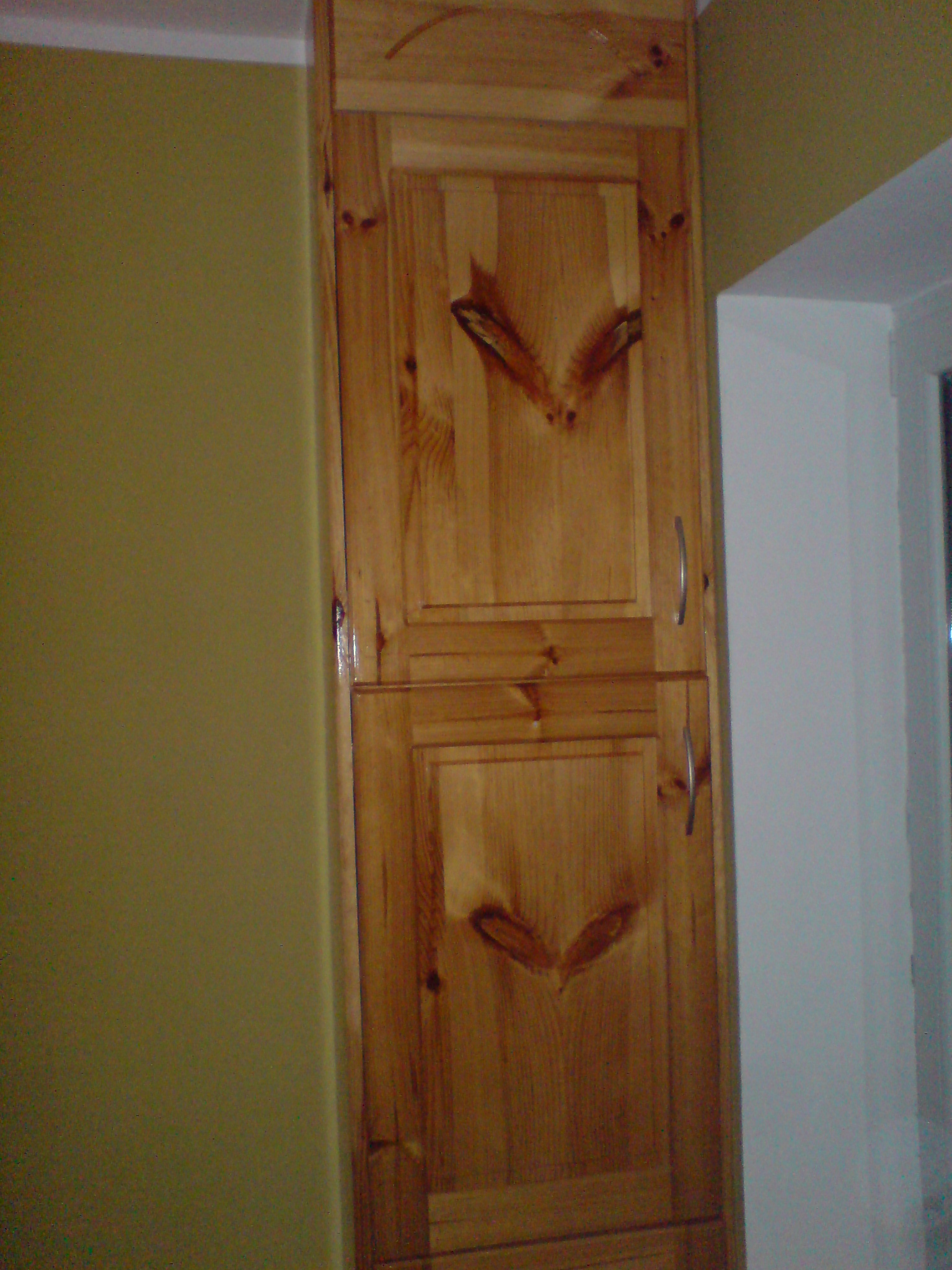 Wysoka, dwudrzwiowa szafa z jasnego drewna sosnowego, z widocznymi sękami i metalowymi uchwytami, stojąca przy ścianie pomalowanej na oliwkowy kolor, obok okna.