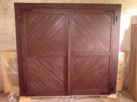 Dwie drewniane okiennice z ukośnym wzorem desek, zawieszone na ciemnych zawiasach, gotowe do montażu.