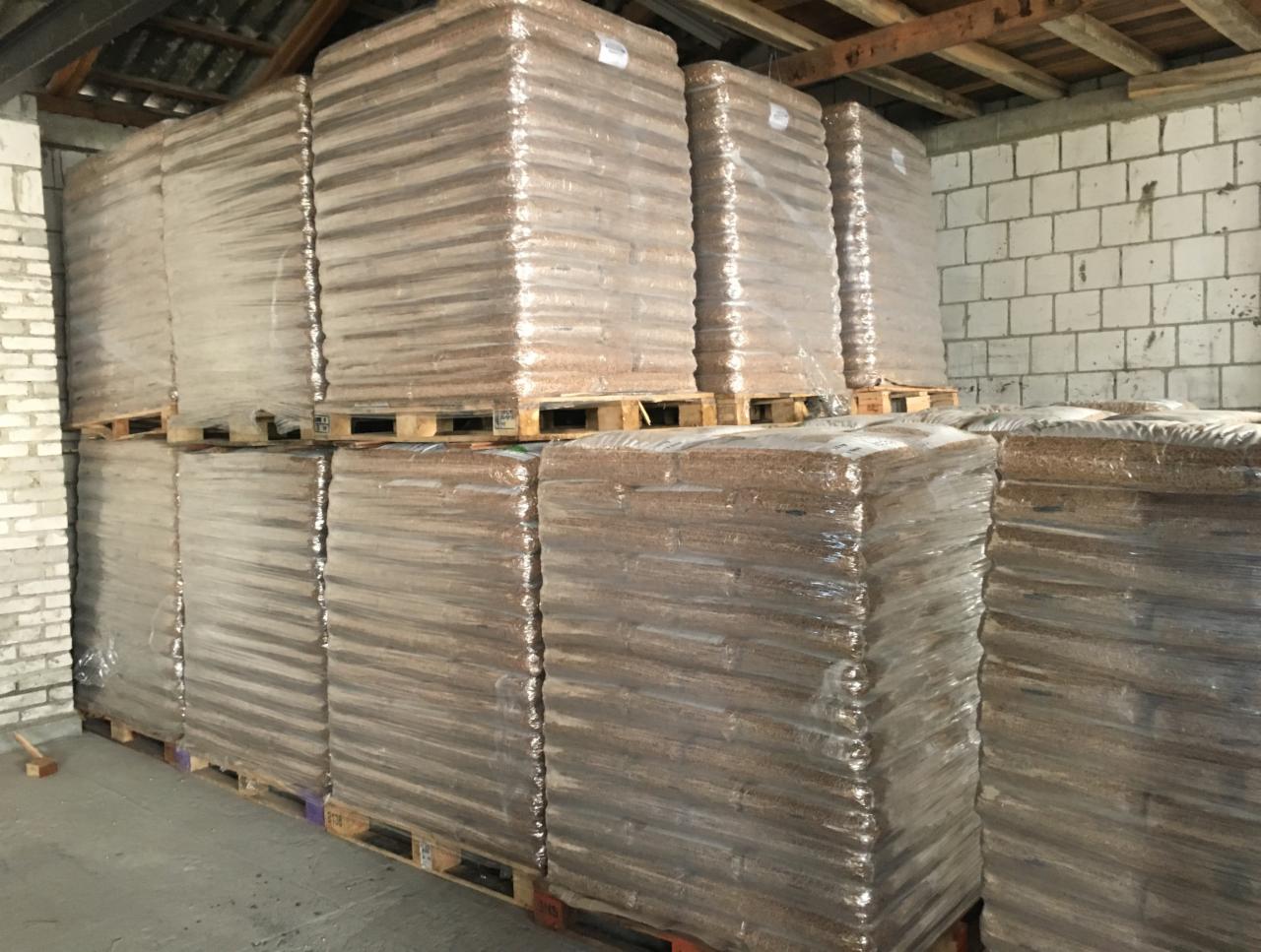 Magazyn: palety z workami pelletu, owinięte folią, ułożone w stosy pod drewnianą konstrukcją dachu, ściany z białego pustaka.