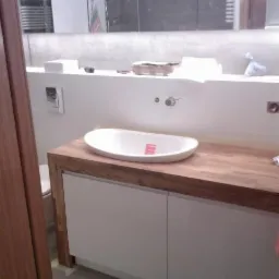 Nowoczesna łazienka w trakcie wykańczania: owalna umywalka nablatowa z różową naklejką na blacie z drewna, białe szafki pod umywalką, szara ściana z dużym lustrem z oświetleniem LED.