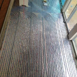 Niebieskie rurki ogrzewania podłogowego ułożone na folii refleksyjnej z czerwoną siatką, widoczne okno i fragment ściany z elementami instalacji.