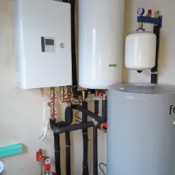 Biała pompa ciepła Toshiba wisząca na ścianie z podłączonymi rurami, obok biały zbiornik buforowy i szary zasobnik ciepłej wody użytkowej, widoczne zawory i manometry.