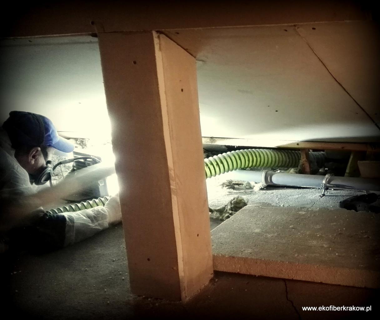 Pracownik w kombinezonie i masce ochronnej natryskuje izolację na poddaszu, widoczny fragment drewnianej konstrukcji i zielony wąż urządzenia.