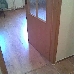 Fragment wnętrza: drewniana podłoga przechodząca w brązowe płytki, widoczna część otwartych drzwi z szybą.