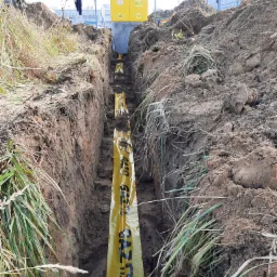 Wykop w ziemi z żółtą taśmą ostrzegawczą 'GAZ 112' wzdłuż rury, widoczna skrzynka gazowa na betonowym fundamencie, ziemia z trawą po bokach wykopu.