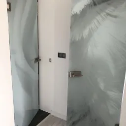 Dwie szklane drzwi z piaskowanymi wzorami: abstrakcyjne fale i motyw piór, z satynowymi okuciami, zamontowane w białym wnętrzu z panelami sterowania oświetleniem.