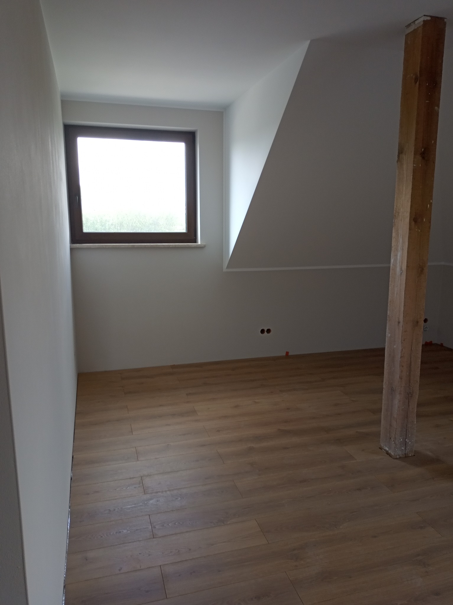 Pokój na poddaszu z drewnianą podłogą i oknem, ściany pomalowane na szaro, widoczny drewniany słup konstrukcyjny.